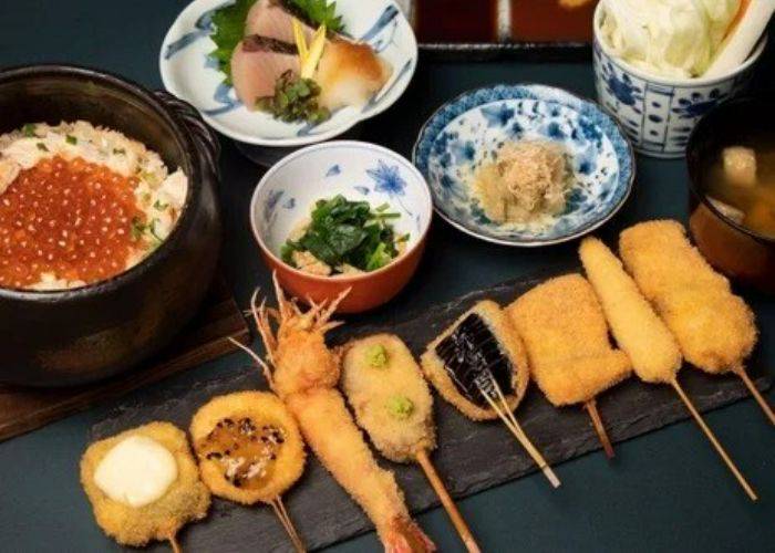 A line of various skewers laid out on a stone serving block, amongst other Japanese dishes.